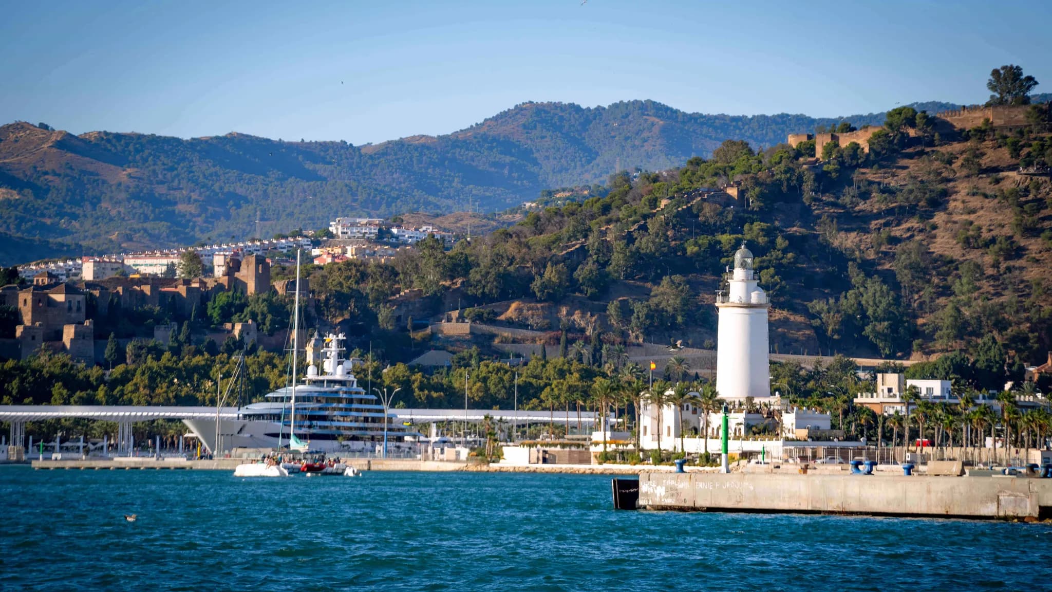 El puerto de Málaga con el faro de La Farola, un superyate en el muelle y la Alcazaba en la ladera arbolada