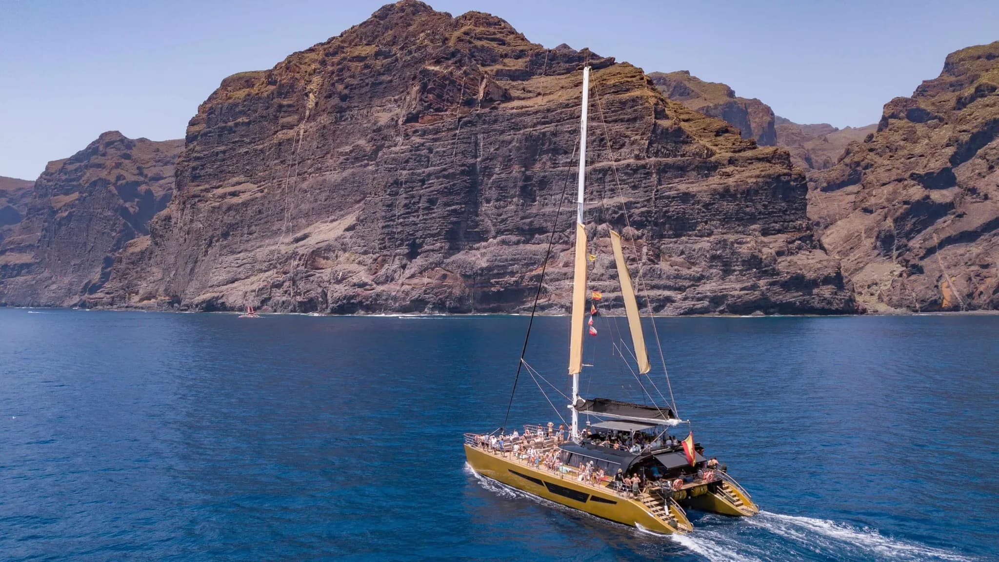 Un catamarán lleno de invitados navegando frente a unos imponentes acantilados
